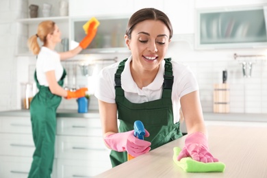 Photo of Woman using rag and sprayer for cleaning table with colleague in kitchen
