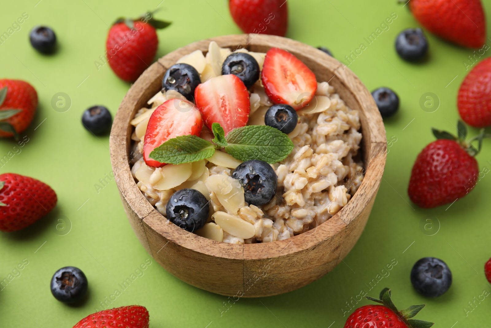 Photo of Tasty oatmeal with strawberries, blueberries and almond petals in bowl surrounded by fresh berries on green background