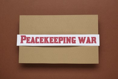 Photo of Paper card with phrase Peacekeeping War on brown background, top view