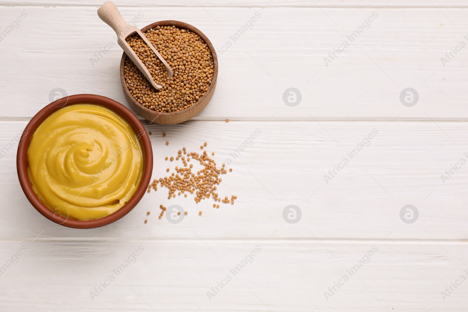 Photo of Bowl with delicious mustard and seeds on white wooden table, flat lay. Space for text