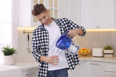 Photo of Happy man pouring water from filter jug into glass in kitchen