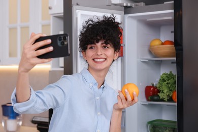 Photo of Smiling food blogger with orange taking selfie near fridge in kitchen
