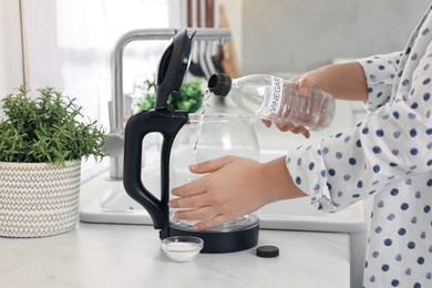 Photo of Woman pouring vinegar from bottle into electric kettle in kitchen, closeup