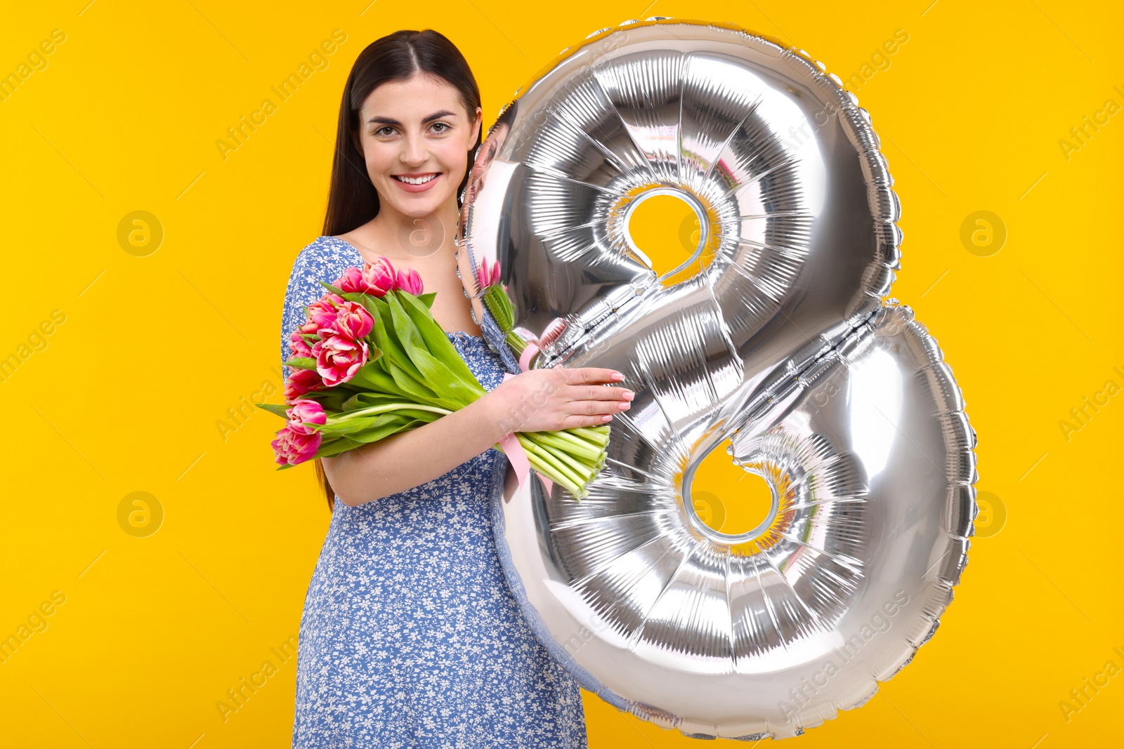 Photo of Happy Women's Day. Charming lady holding bouquet of beautiful flowers and balloon in shape of number 8 on orange background