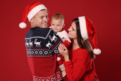 Photo of Happy couple with cute baby in Christmas outfits and Santa hats on red background