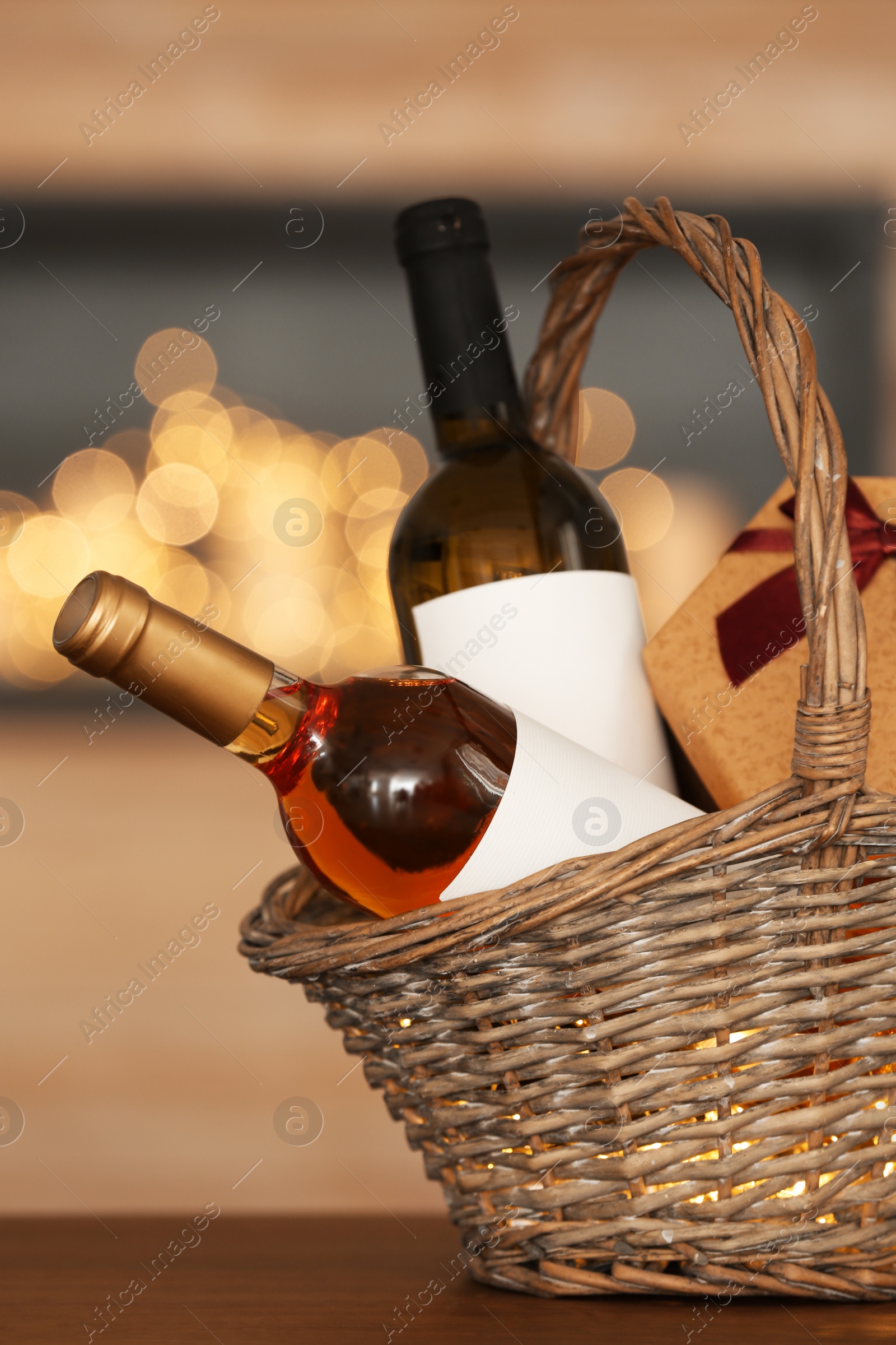 Photo of Wicker basket with bottles of wine and gift box on table against blurred background