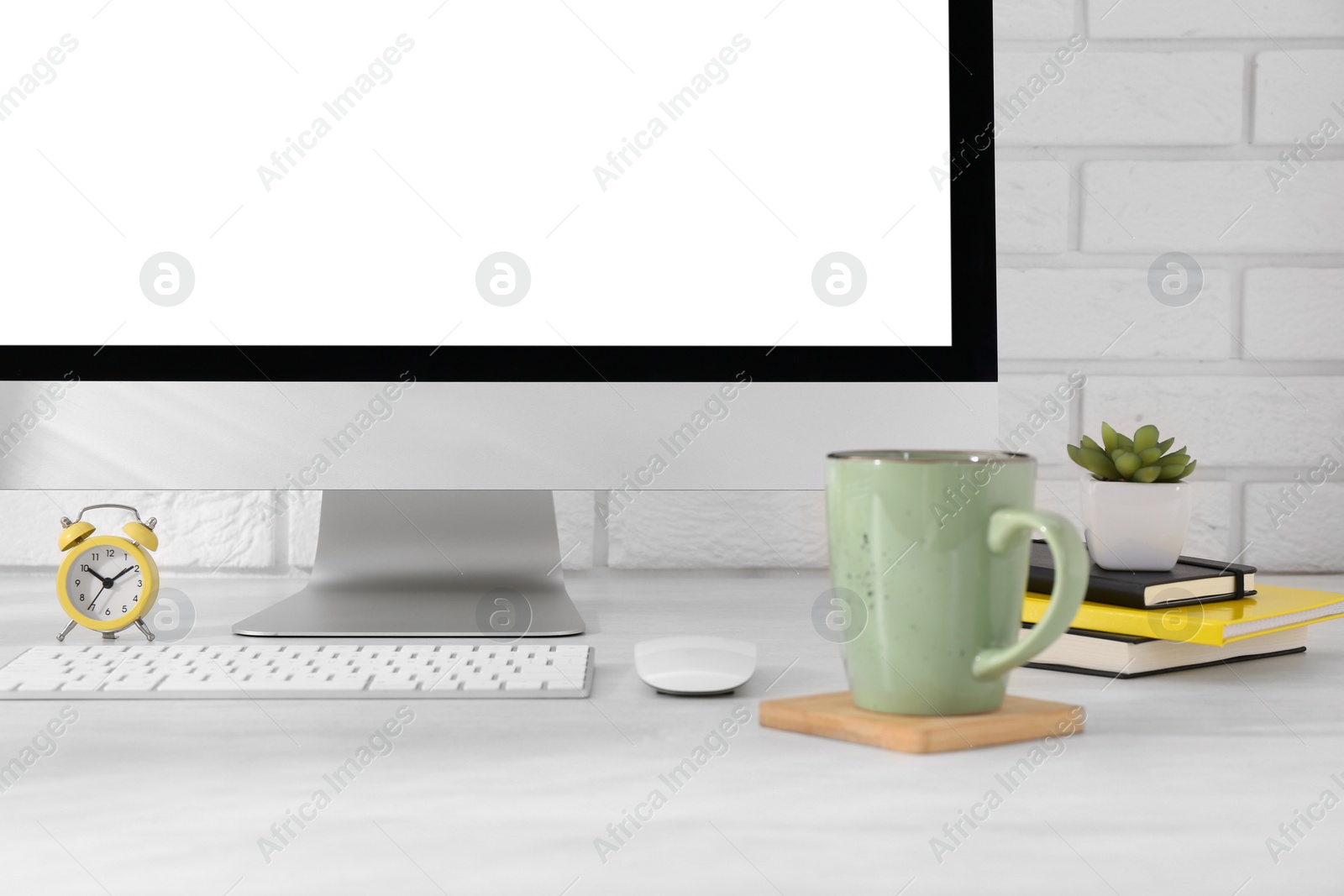 Photo of Office workplace with computer, stationery, cup, alarm clock and houseplant on light table near white brick wall