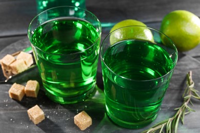 Photo of Absinthe in glasses, rosemary, brown sugar and lime on black wooden table, closeup. Alcoholic drink