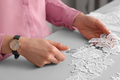 Photo of Dressmaker cutting beautiful white lace at table in atelier, closeup