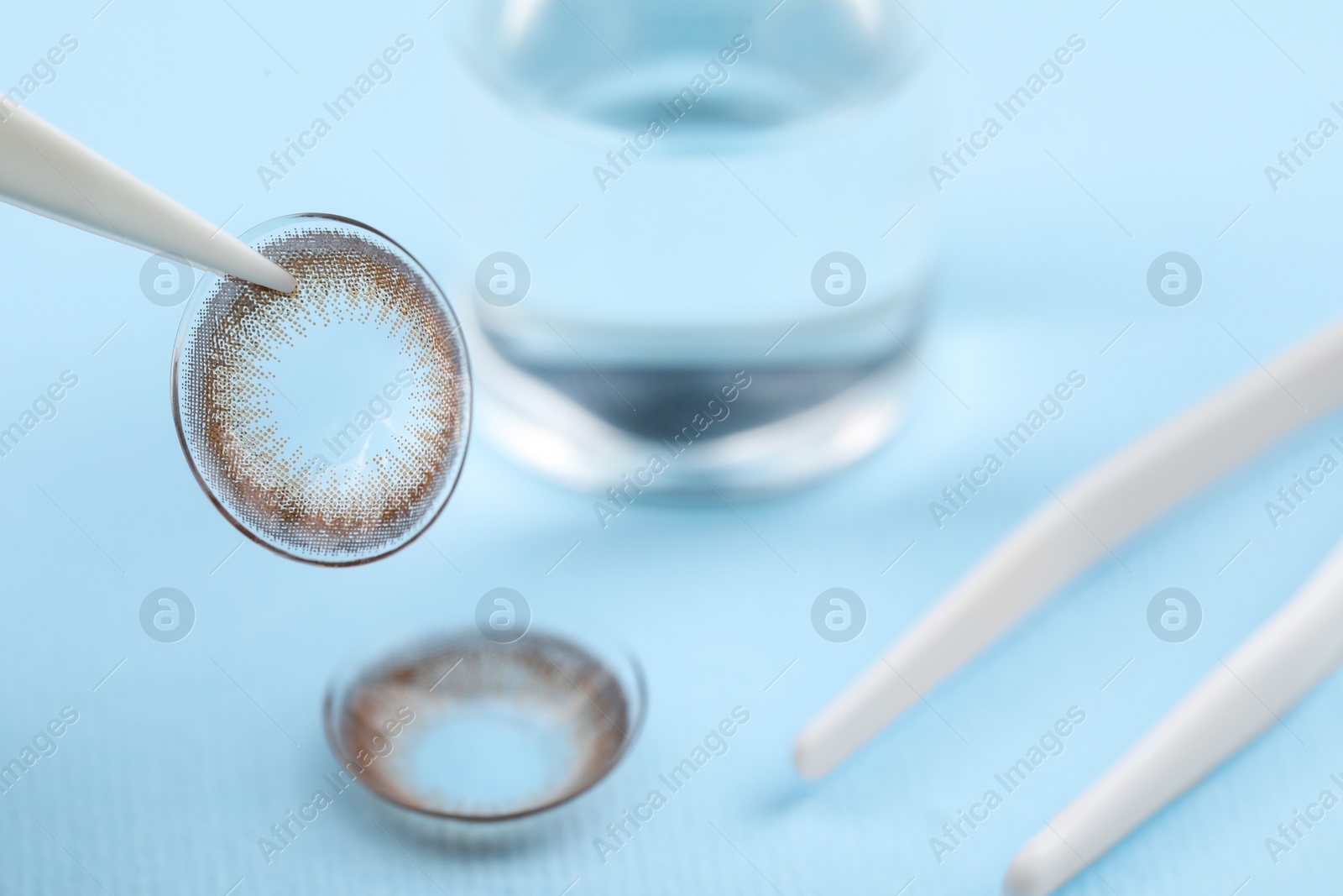 Photo of Tweezers with color contact lens on light blue background, closeup. Space for text