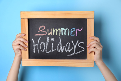 Photo of Schoolgirl holding blackboard with text SUMMER HOLIDAYS on light blue background, closeup