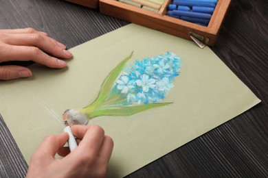 Photo of Woman drawing blue blooming hyacinth on paper with soft pastels at wooden table, closeup