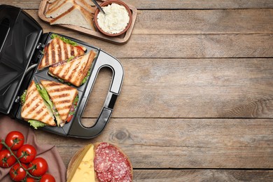 Photo of Modern grill maker with sandwiches and different products on wooden table, flat lay. Space for text