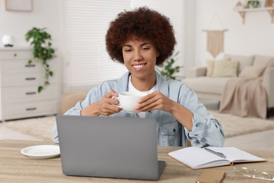 Photo of Young woman using laptop and drinking coffee at wooden desk in room