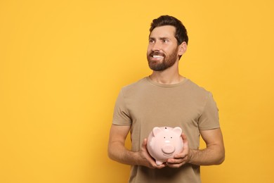 Photo of Happy man with ceramic piggy bank on orange background, space for text