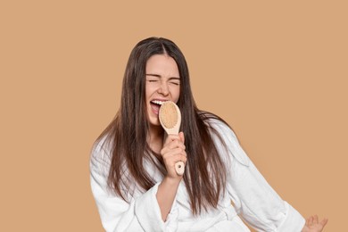 Photo of Emotional woman in bathrobe singing into brush on beige background. Spa treatment