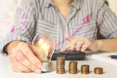 Image of Woman with light bulb, calculator and coins at white table, closeup. Energy saving