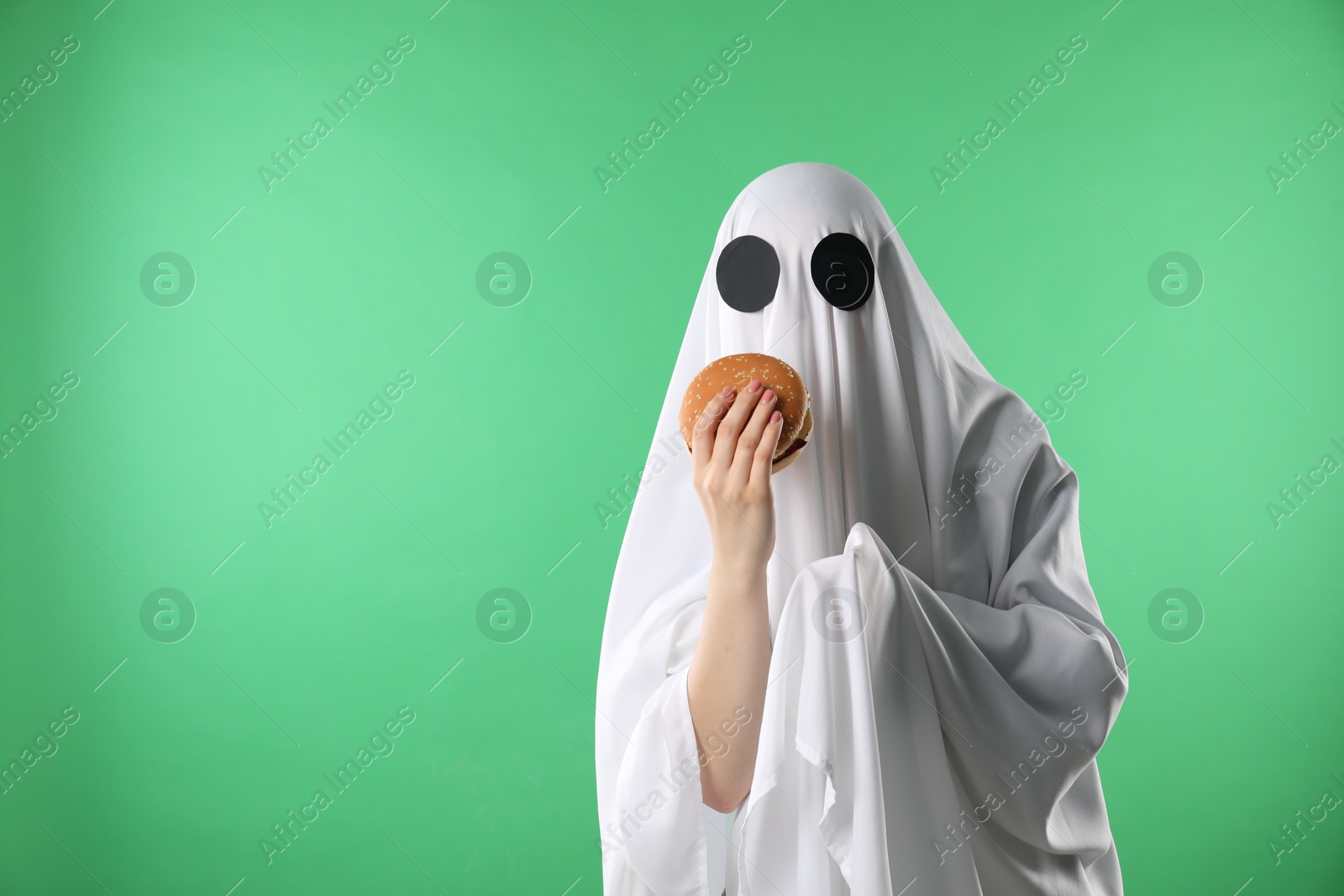 Photo of Creepy ghost. Person in white sheet with tasty hamburger on green background, space for text