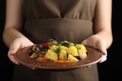 Woman holding earthenware with delicious roasted potatoes and meat on black background, closeup