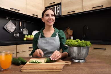 Photo of Woman in apron cutting cucumber at wooden table indoors