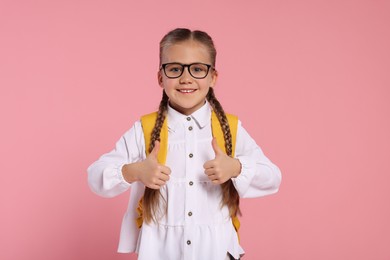 Happy schoolgirl with backpack showing thumbs up gesture on pink background