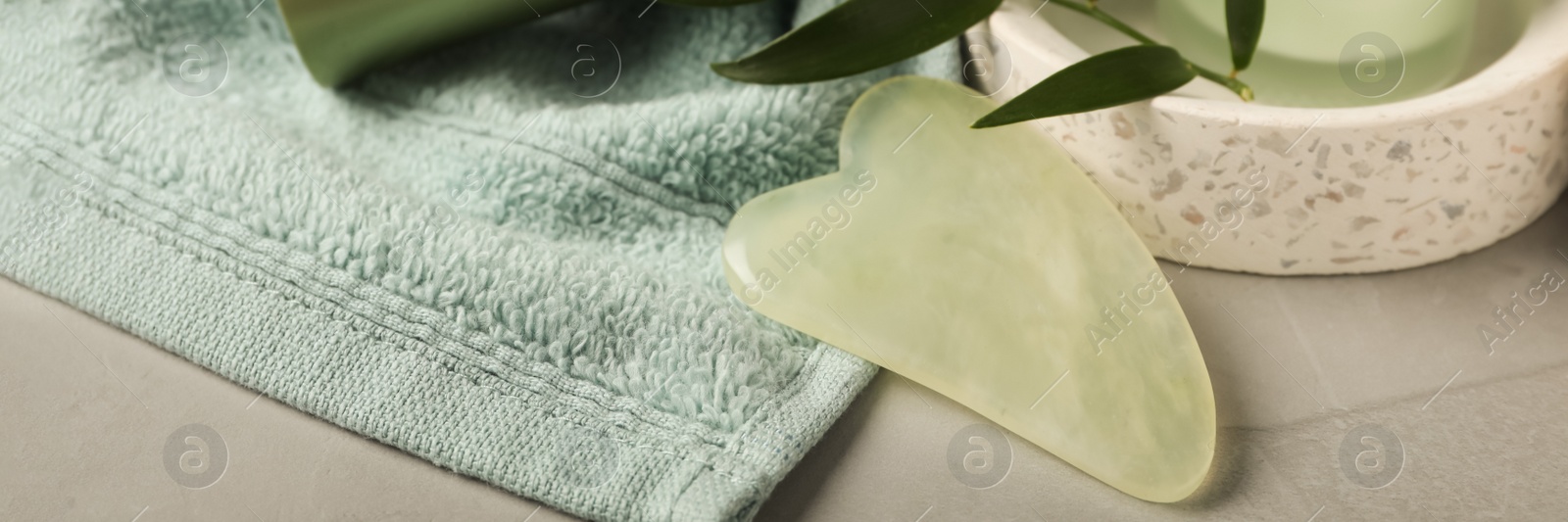 Image of Jade gua sha tool, skin care products and towel on grey table. Banner design