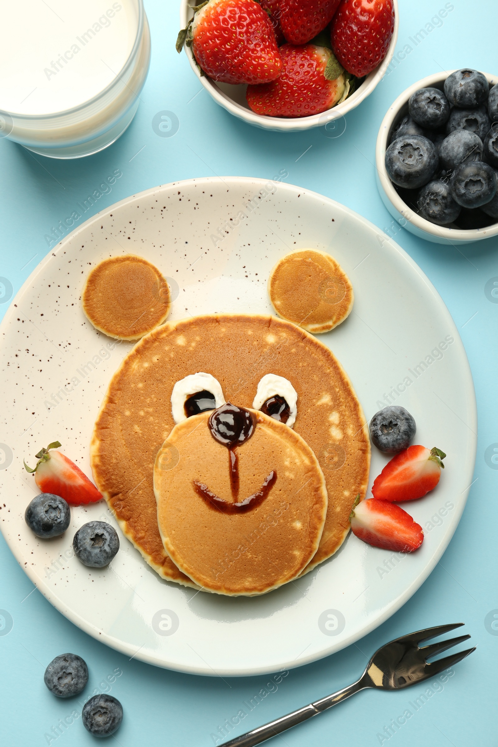 Photo of Creative serving for kids. Plate with cute bear made of pancakes and berries on light blue table, flat lay