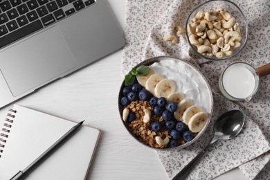 Photo of Delicious granola in bowl, stationery and laptop on white wooden table, flat lay