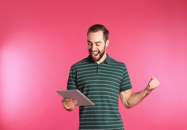 Emotional young man with tablet celebrating victory on color background