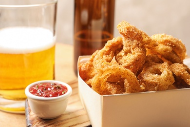 Photo of Cardboard box with crunchy fried onion rings and tomato sauce on wooden board, closeup