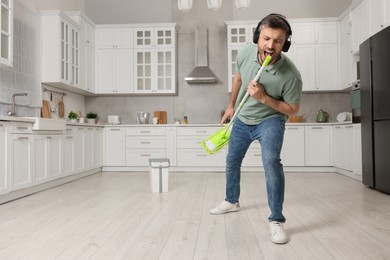 Photo of Happy man in headphones with mop singing while cleaning in kitchen. Space for text