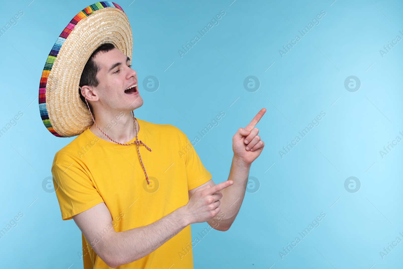 Photo of Young man in Mexican sombrero hat pointing at something on light blue background. Space for text