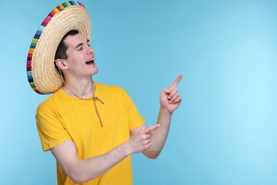 Photo of Young man in Mexican sombrero hat pointing at something on light blue background. Space for text