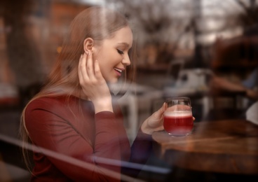 Pretty young woman with cocktail at table in cafe, view from outdoors through window