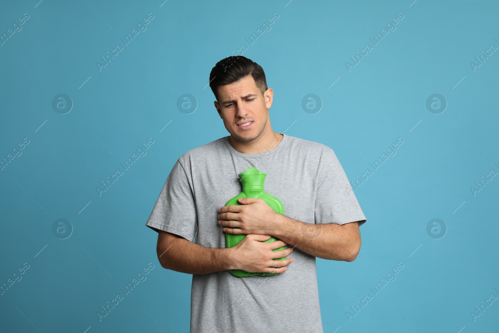 Photo of Man using hot water bottle to relieve chest pain on light blue background