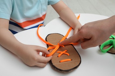 Photo of Mother teaching son to tie shoe laces using training cardboard template at white table, closeup