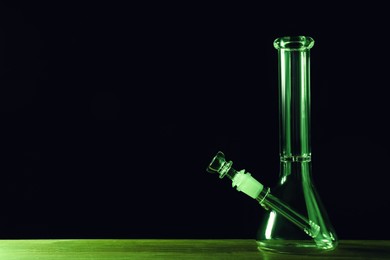 Photo of Glass bong on wooden table against black background, space for text. Smoking device