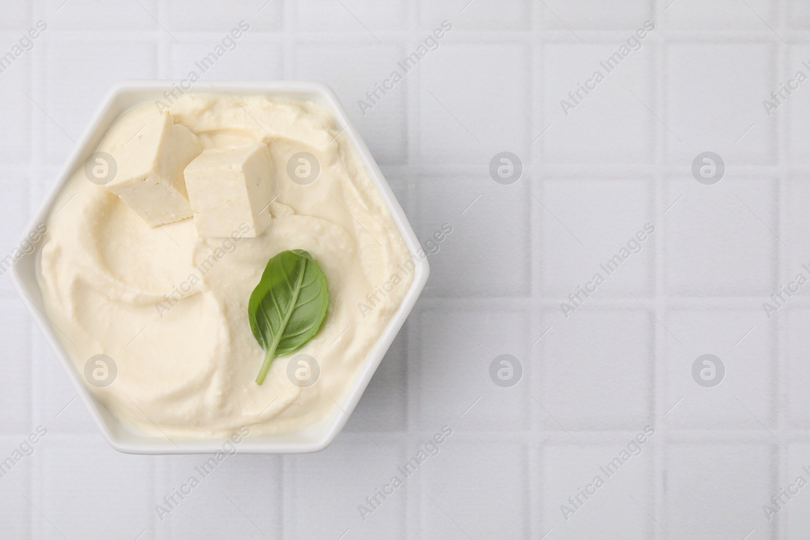 Photo of Delicious tofu sauce and basil leaf in bowl on white tiled table, top view. Space for text