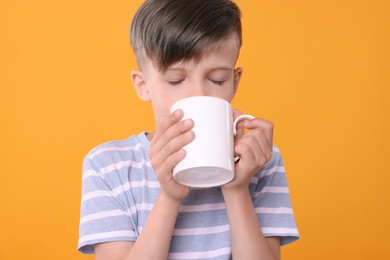 Photo of Cute boy drinking beverage from white ceramic mug on orange background