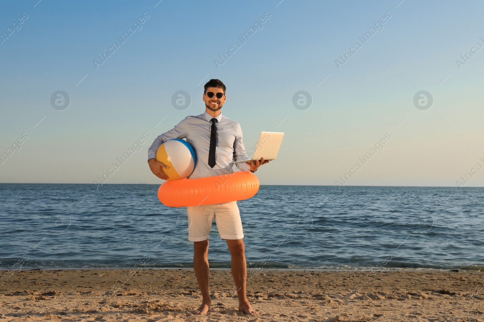 Photo of Happy man with inflatable ring, ball and laptop near sea on beach. Business trip