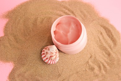 Open jar of cream and seashell on sand against pink background, above view