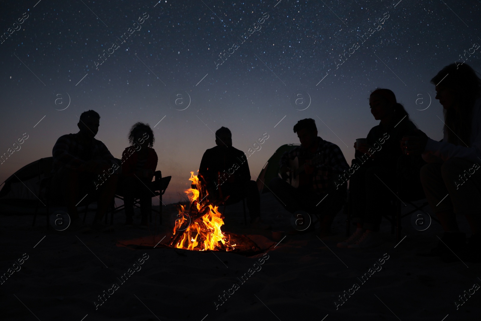 Photo of Group of friends gathering around bonfire in evening. Camping season
