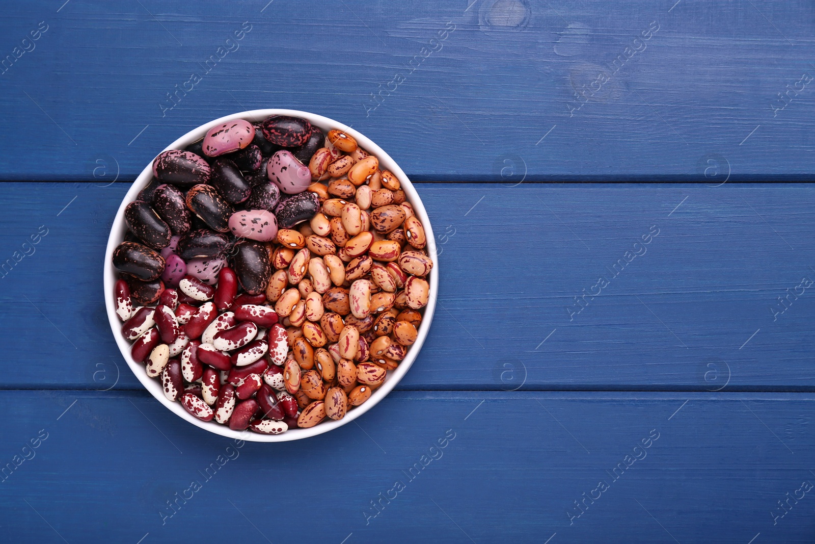 Photo of Different kinds of dry kidney beans in bowl on blue wooden table, top view. Space for text