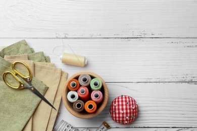 Flat lay composition with spools of threads and sewing tools on white wooden table, space for text
