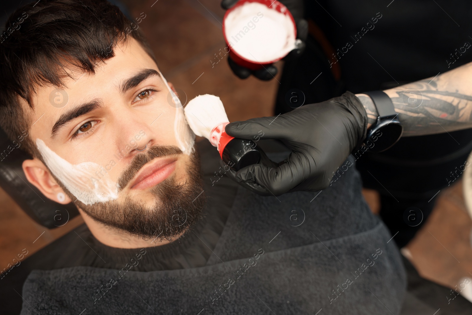 Photo of Professional hairdresser applying shaving foam onto client's beard in barbershop, closeup