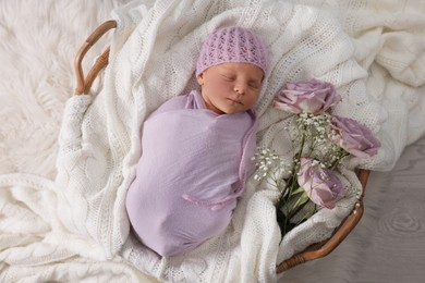 Photo of Adorable newborn baby lying in basket with knitted plaid and flowers on floor, top view