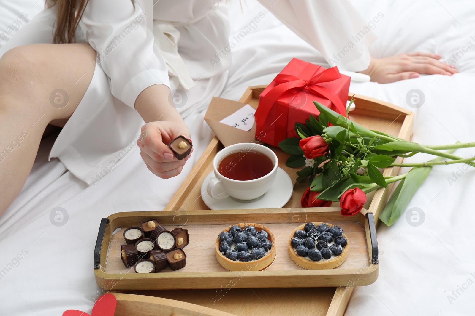 Photo of Tasty breakfast served in bed. Woman with desserts, tea, gift box and flowers at home, closeup