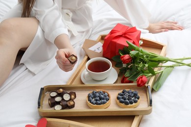 Photo of Tasty breakfast served in bed. Woman with desserts, tea, gift box and flowers at home, closeup