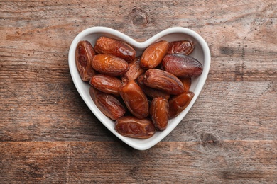 Photo of Heart shaped bowl with sweet dried date fruits on wooden background, top view