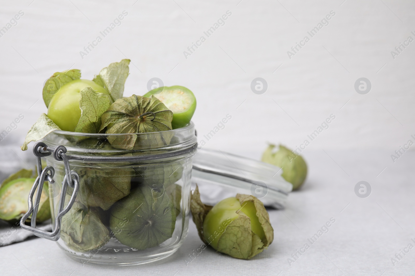 Photo of Fresh green tomatillos with husk in glass jar on light table, space for text